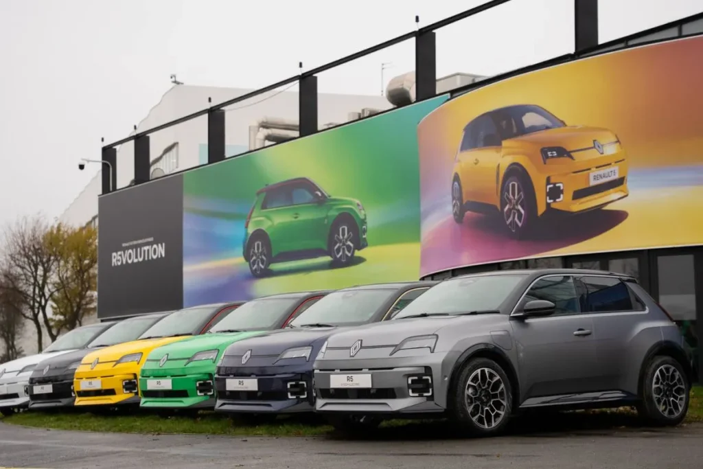 Que carros elétricos têm direito ao incentivo à compra? 5 Renault 5 de várias cores, estaconados frente a um cartaz. O R5 enquadra-se no incentivo à compra